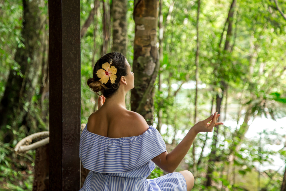 A woman in a blue off-shoulder dress sits in meditation at an Ayahuasca retreat in Colombia, surrounded by lush green jungle, embracing a peaceful and spiritual experience.
