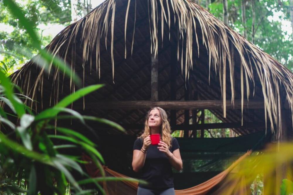 Woman holding a red cup and standing in front of a thatched jungle hut, symbolizing the immersive natural setting of Peyote Retreats in Costa Rica. 