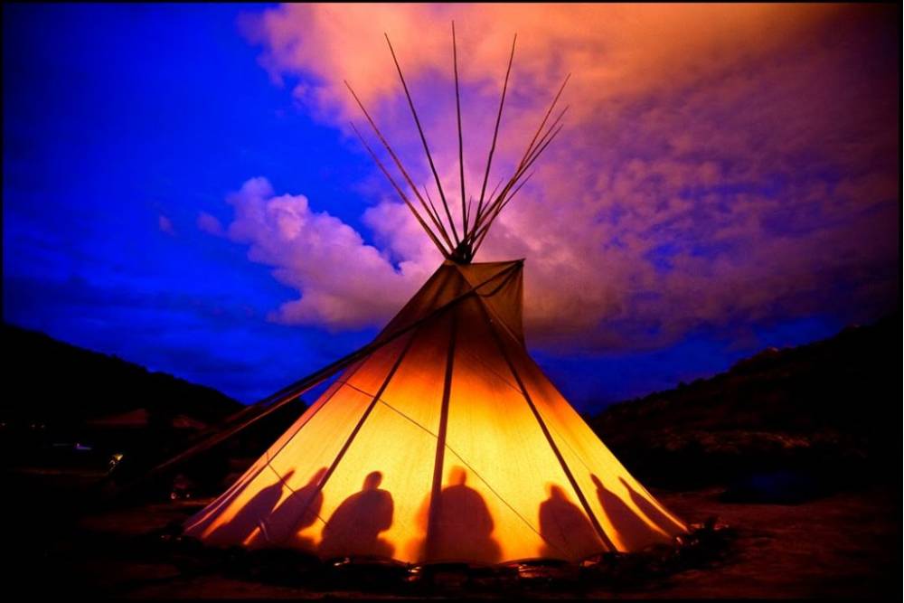 A glowing tipi at dusk with silhouettes of people inside, symbolizing a sacred gathering space for those asking where can I do a peyote ceremony.