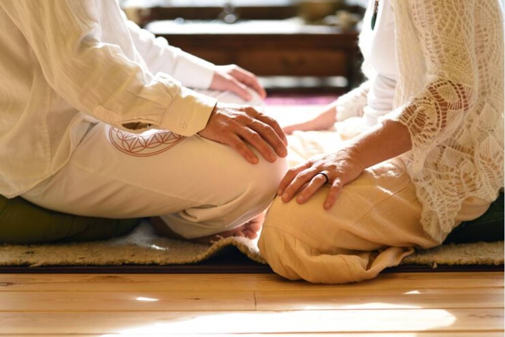 Two individuals sitting in ceremonial posture with gentle physical connection, representing the intimate healing process of an iboga ceremony.